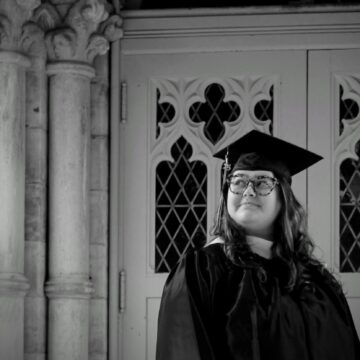 Nagel stands in front of chapel doors in a graduation cap and gown, looking off to the side with quiet pride. The black-and-white photo symbolizes the bittersweet nature of their college experience and the strength it took to fight for gender justice on campus.
