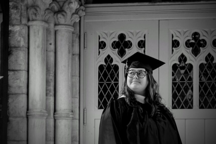 Nagel stands in front of chapel doors in a graduation cap and gown, looking off to the side with quiet pride. The black-and-white photo symbolizes the bittersweet nature of their college experience and the strength it took to fight for gender justice on campus.