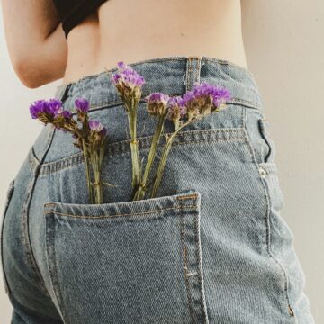 Close-up of a person wearing high-waisted denim jeans with purple flowers tucked into the back pocket. The photo symbolizes strength, softness, and survival, reflecting the spirit of Denim Day and the fight against sexual violence.