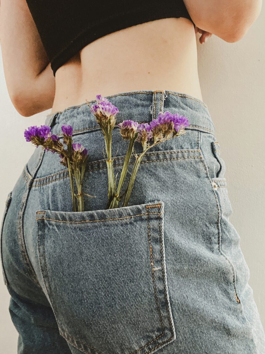 Close-up of a person wearing high-waisted denim jeans with purple flowers tucked into the back pocket. The photo symbolizes strength, softness, and survival, reflecting the spirit of Denim Day and the fight against sexual violence.