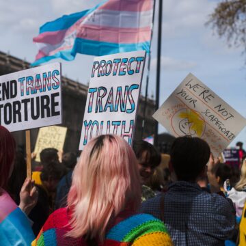 a transgender rights protest. people hold signs depicting the trans flag with sayings like "protect trans youth" and "end trans torture"