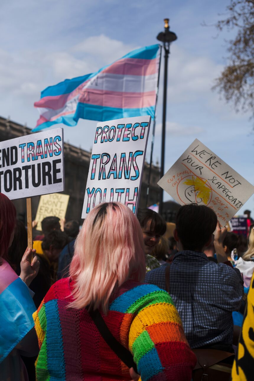 a transgender rights protest. people hold signs depicting the trans flag with sayings like "protect trans youth" and "end trans torture"