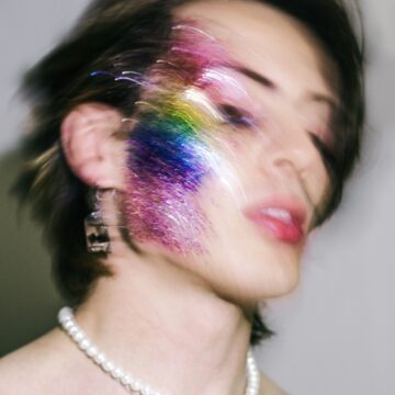 Editorial-style portrait of a person in glittery makeup embodying experimental LGBTQ+ fashion and camp style. The image uses long exposure to highlight vibrant color, movement, and euphoria. With dramatic lighting and theatrical makeup, the photo celebrates gay pride and gender performance, blending film aesthetics, party culture, and non-normative style in a glamorous, high-energy scene.