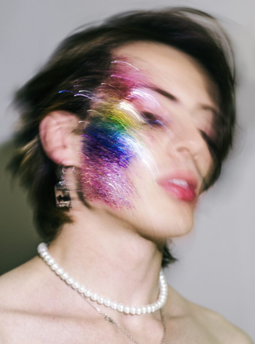 Editorial-style portrait of a person in glittery makeup embodying experimental LGBTQ+ fashion and camp style. The image uses long exposure to highlight vibrant color, movement, and euphoria. With dramatic lighting and theatrical makeup, the photo celebrates gay pride and gender performance, blending film aesthetics, party culture, and non-normative style in a glamorous, high-energy scene.