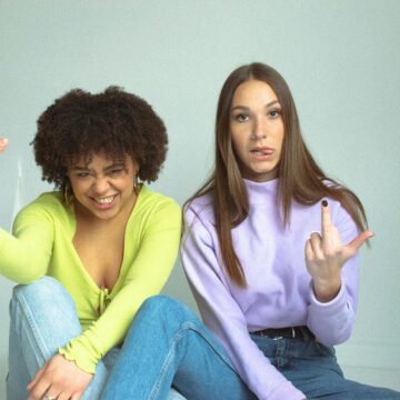 An image of two women flipping off the camera. The woman on the left is in jeans and a greenish yellow long sleeve shirt. The woman on the right is in jeans and a pastel purple turtleneck. Both are sitting on the ground with an off-white backdrop