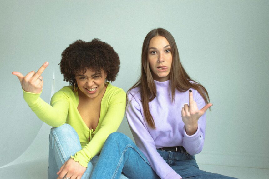An image of two women flipping off the camera. The woman on the left is in jeans and a greenish yellow long sleeve shirt. The woman on the right is in jeans and a pastel purple turtleneck. Both are sitting on the ground with an off-white backdrop