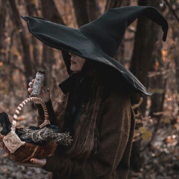 A photo of a modern witch in a black witch hate, holding a basket and a bundle of sage