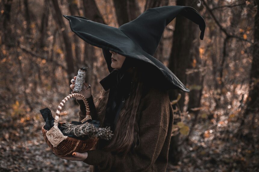 A photo of a modern witch in a black witch hate, holding a basket and a bundle of sage