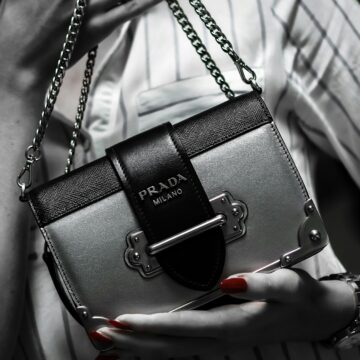 A black and white image of a woman holding a Prada purse. The only part of this image that is in color is her red nails. She is wearing a striped shirt and holding the purse in frame, while her face is out of frame, adding to the theme of materialism that is prevalent in this post.