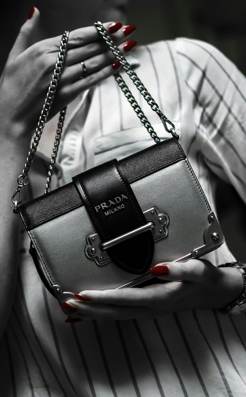 A black and white image of a woman holding a Prada purse. The only part of this image that is in color is her red nails. She is wearing a striped shirt and holding the purse in frame, while her face is out of frame, adding to the theme of materialism that is prevalent in this post.