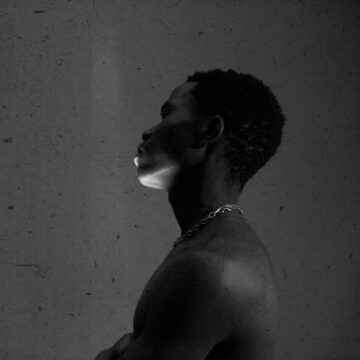 olu-famule-hl_tbLIqQIA-unsplash black and white photo of a Nigerian man standing in a dark room with a cement wall behind him. He's facing to the left and has a streak of light across his face