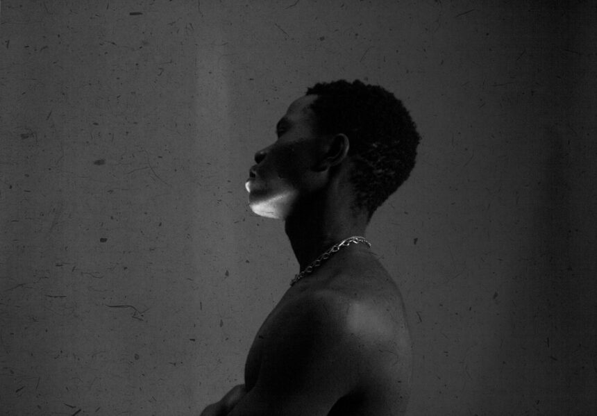 olu-famule-hl_tbLIqQIA-unsplash black and white photo of a Nigerian man standing in a dark room with a cement wall behind him. He's facing to the left and has a streak of light across his face